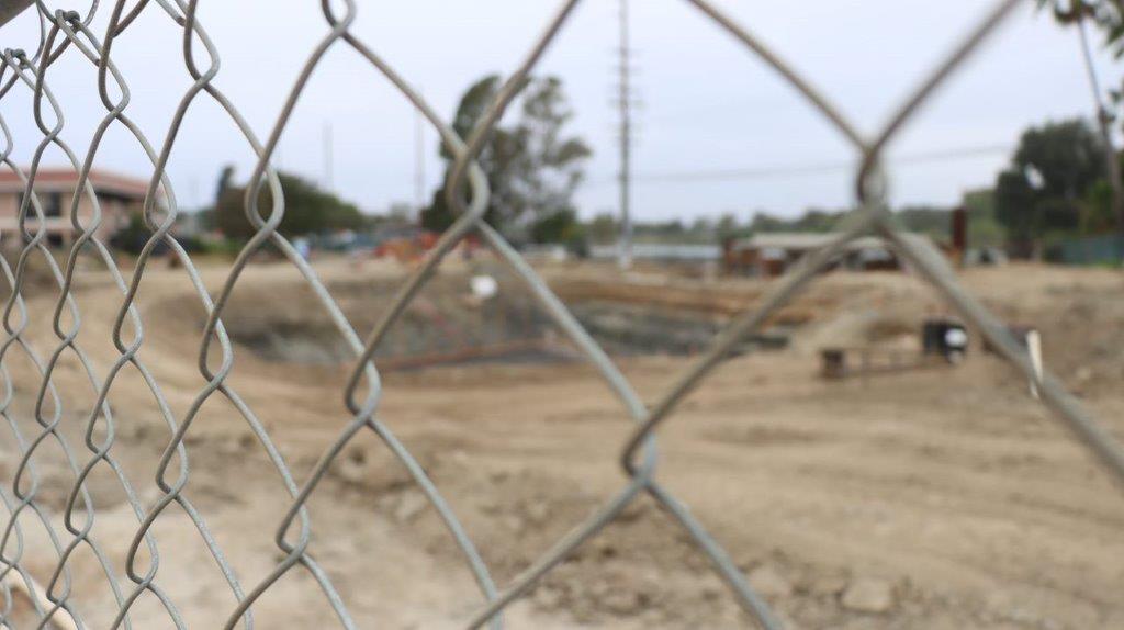 Close-up of a fence with dirt and a building in the background

Description automatically generated